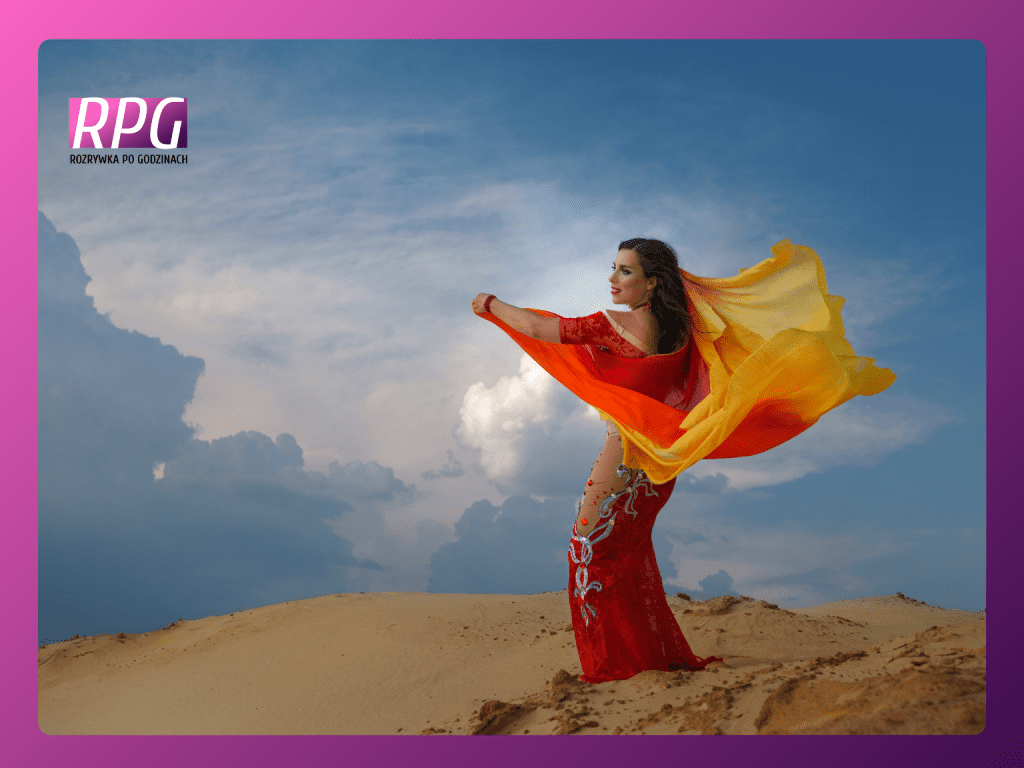 Kobieta stojąca na pustyni, za nią niebo. Kobieta ma czarne włosy i śniadą cerę - jest indyjskiego pochodzenia. Ubrana jest w czerwono pomarańczowe sari, powiewająca na wietrze